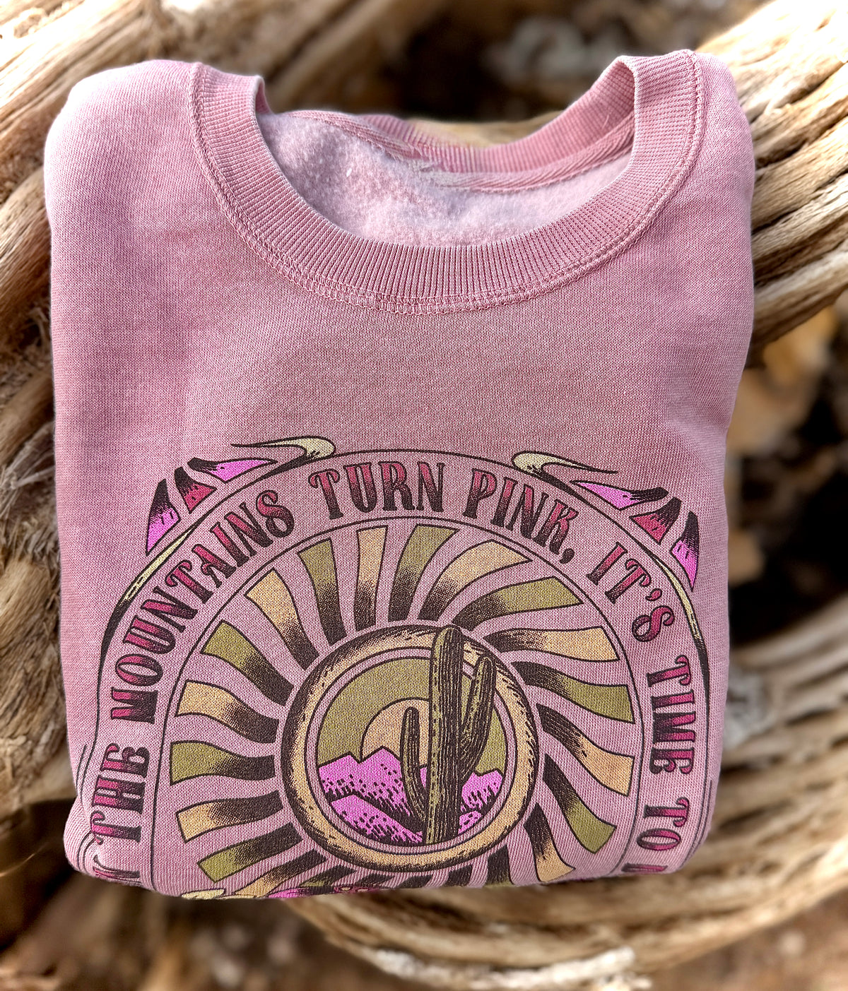 Faded pink crewneck sweatshirt featuring a retro desert sun graphic with cactus, mountains, and the quote The Mountains Turn Pink.