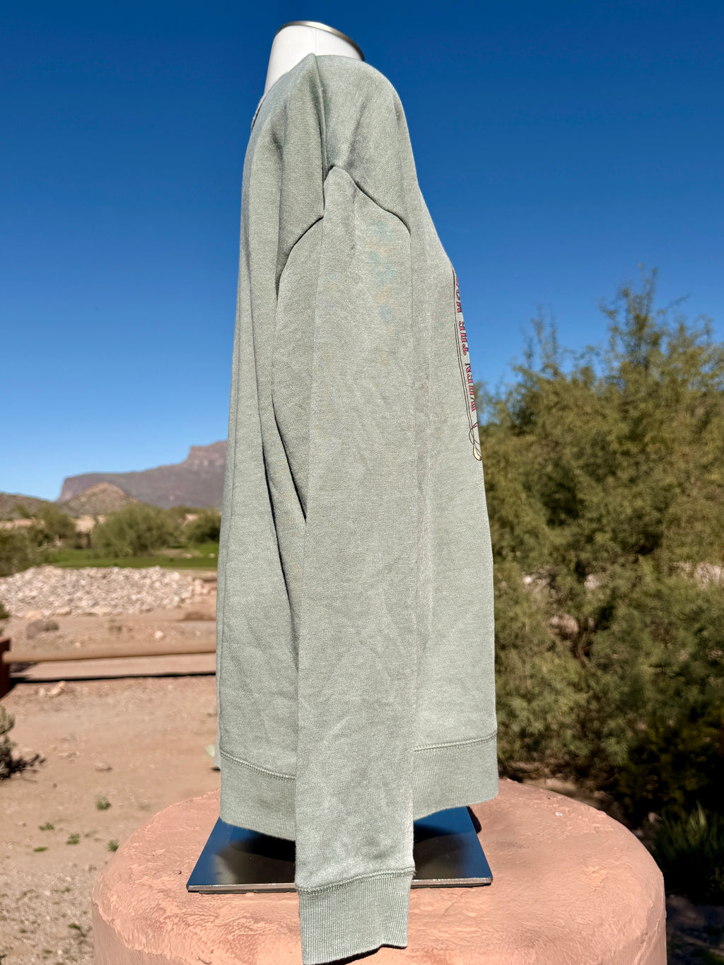 Side view of a weathered sage green crewneck sweatshirt with a vertical graphic, displayed outdoors against a blue sky and desert mountains.