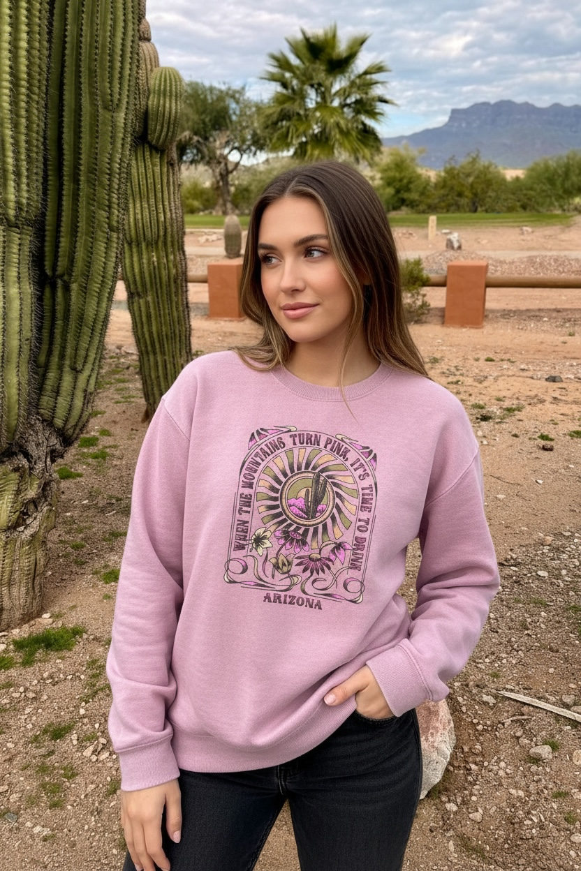 Pink crewneck sweatshirt featuring retro desert cactus graphic and "When The Mountains Turn Pink" text, worn near a Saguaro in Arizona.